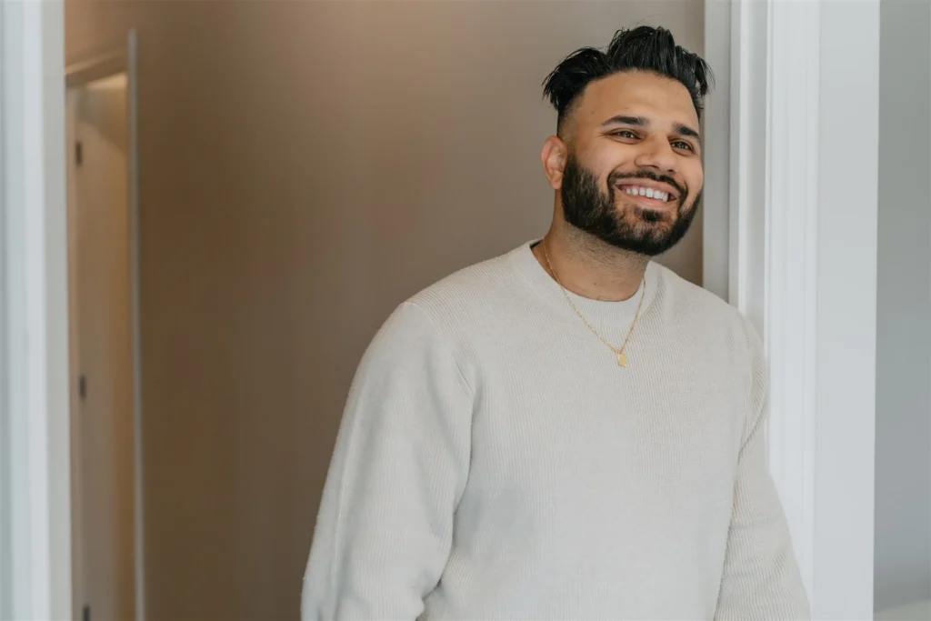 A man in a beige sweater smiles in an open doorway.