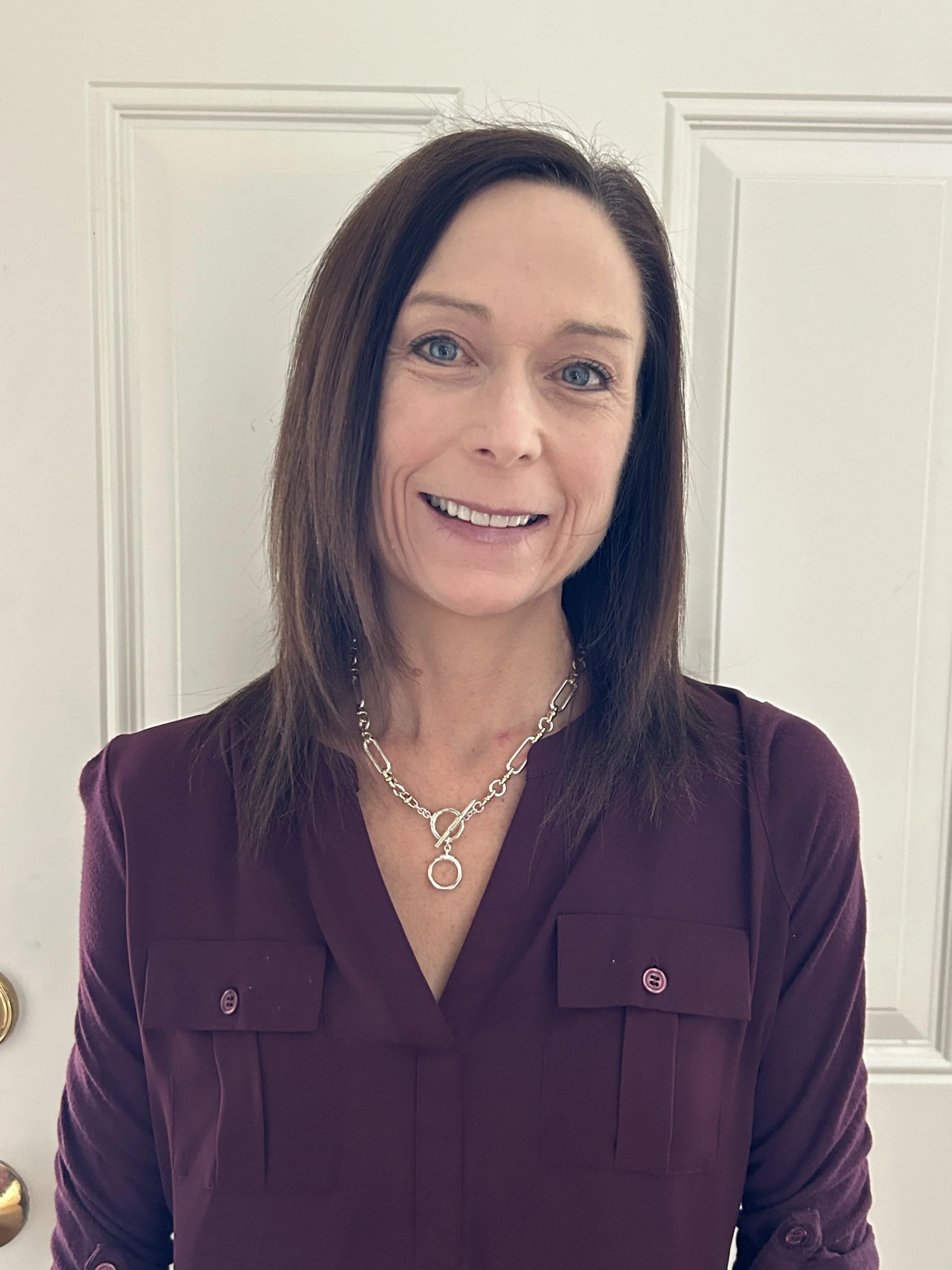 Portrait of a smiling woman with long brown hair wearing a burgundy blouse and a silver necklace.