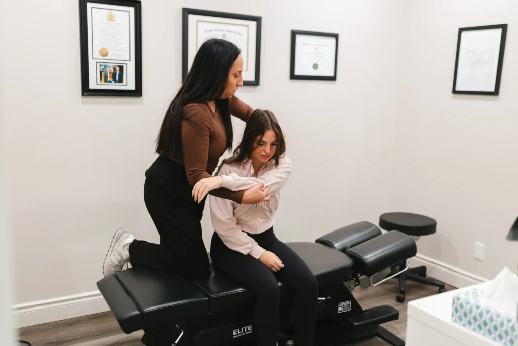 A woman is adjusting another woman's back on a chiropractic table in a clinical setting with framed certificates on the wall.