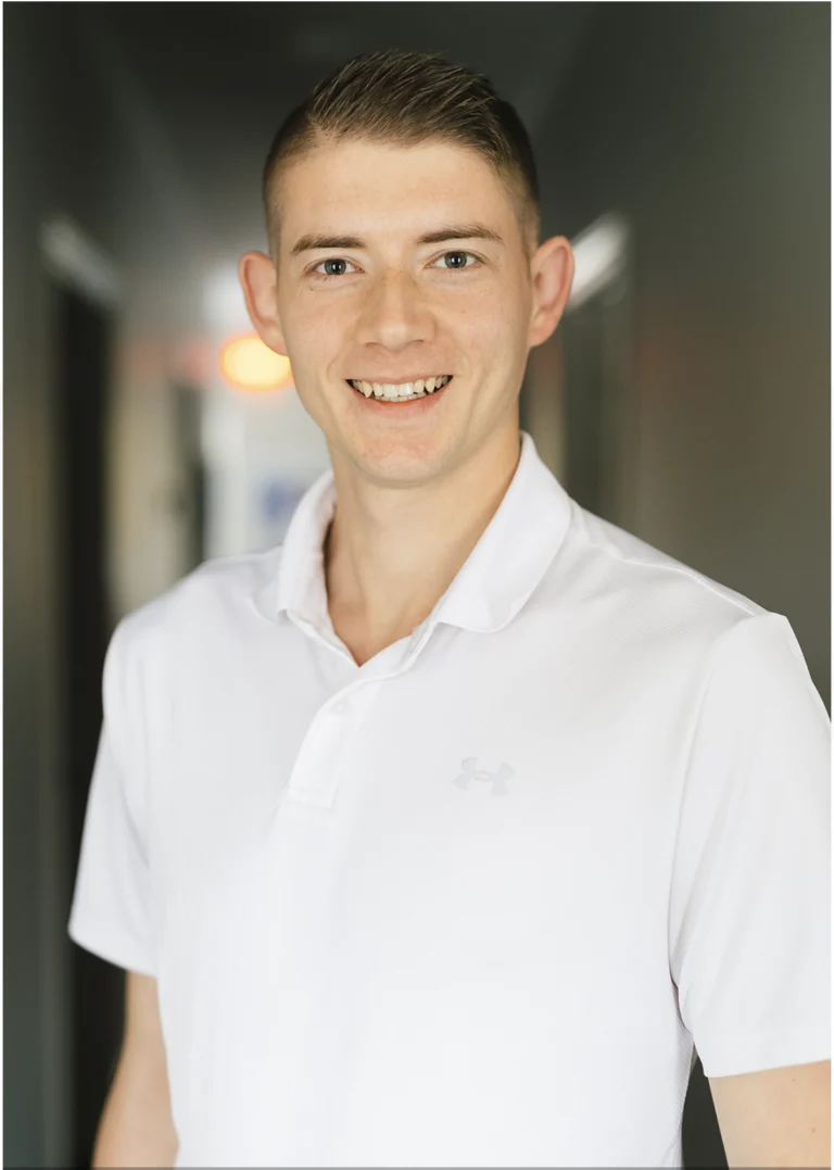 A man in a white collared shirt stands indoors, smiling at the camera with short hair and a blurred hallway background.