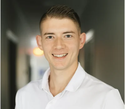 A young man with short hair wearing a white Under Armour polo shirt stands smiling in a hallway with dark walls.